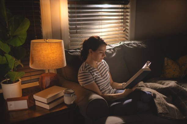 istock Young woman sitting on a cosy sofa and reading a book in low light. 1253344272
