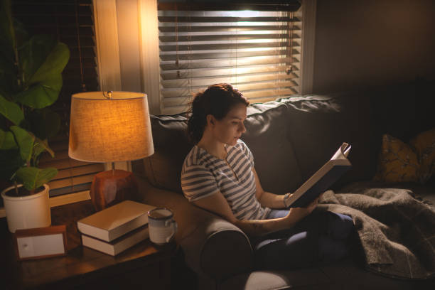 Młoda kobieta siedząca na przytulnej kanapie i czytając książkę w słabym świetle. – zdjęcie