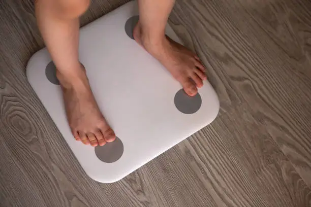Photo of Kid girl or boy stands and weighs himself on white modern electronic smart scale. Scales stand on grey wooden floor. Only legs are visible. Concept of Internet of things, weight control, health