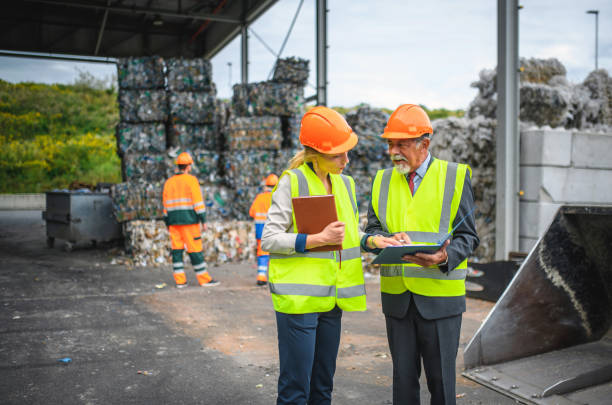 Quality Control Inspectors at Waste Management Facility Male and female quality control inspectors in discussion at waste management facility with workers and stacks of bundled paper and plastics in background. recycling center stock pictures, royalty-free photos & images