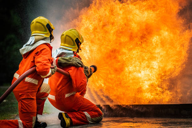 istock Two brave firefighter in fire suit on rescue duty using water from hose extinguishing fighting with big crackle fire flames inside burning premises. Fireman spraying high pressure water fight a fire. 1253392107