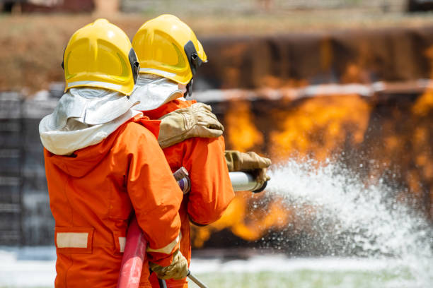 Two Firefighter teamwork in fire suit with fire fighting equipment using high pressure water fight a fire dos valientes bomberos en traje de fuego en servicio de rescate utilizando agua de la extinción de mangueras luchando con grandes llamas de fuego crepitante dentro de las instalaciones en llamas. el bombero rociando agua a alta presión combate un incendi