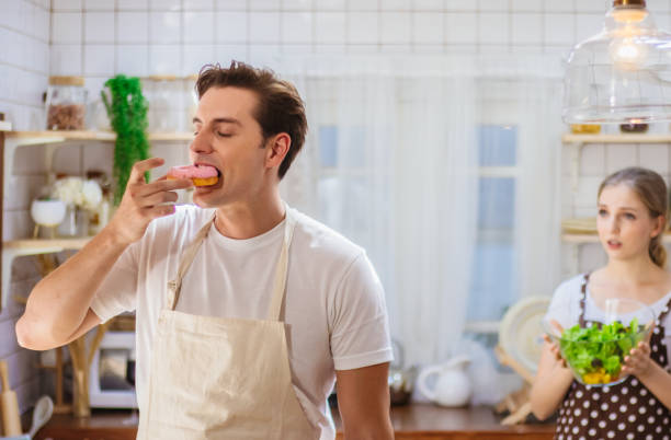 beau couple caucasien ou amant dans la cuisine ensemble. l’homme heureux mangent la malbouffe malsaine comme donut tandis que la salade de cuisson de femme pour le repas. petite amie malheureuse bouleversée ou déçue sur de mauvaises habitudes de petit - overweight women salad frustration photos et images de collection