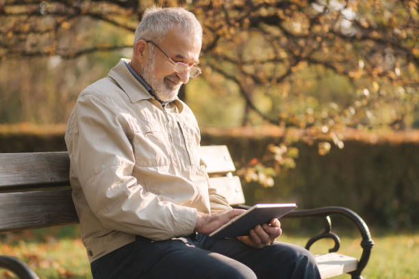 istock Handsome elderly man sitting on the bench and use tablet for scrolling in internet. Background of autumn tree 1253429291