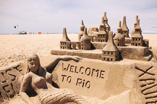 Copacabana, Rio de Janeiro, Brazil - June 12, 2016: View of the symbolic sidewalk of Copacabana with people, a sand castle, a Brazilian flag, the beach and the structures of the future Olympic beach volleyball complex.