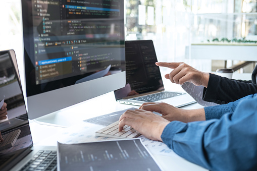 A person points at code displayed on a large monitor while another person types on a keyboard in front of a laptop, with design documents spread across the table in a modern workspace.