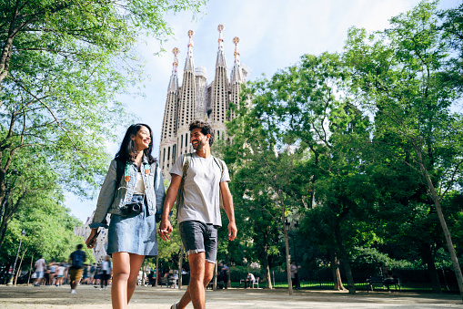Low angle view of smiling vacationers holding hands and walking through lush green Barcelona park with Sagrada Familia in background.