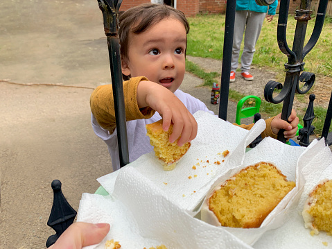 A close up of a young mixed race boy standing on his driveway reaching his hand out through the iron gates to grab a piece of cake from a plate that an unrecognizable person is holding.