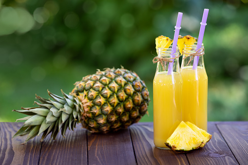 pineapple juice in glass bottles and ripe fruit on wooden table outdoors. Summer refreshing drink