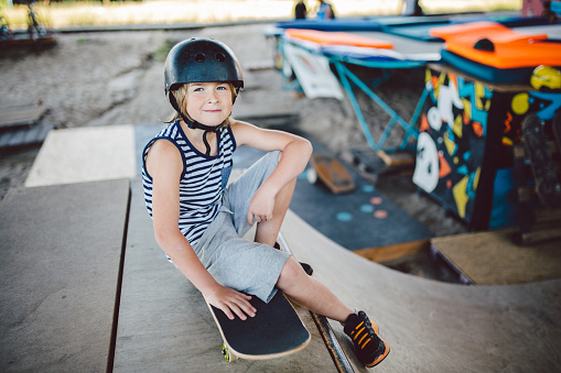 Boy sitting at skatepark, looking camera. Kid resting with skate board at skate park. child sits on ramp resting after training on skateboard. Trendy youngster enjoying free time at half pipe ramp.