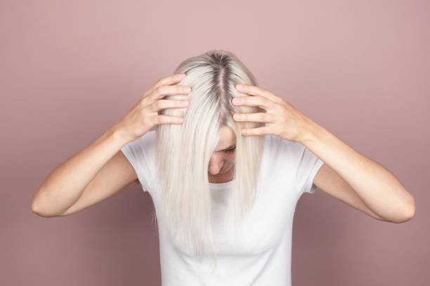 hermosa joven rubia en una camiseta blanca muestra raíces de pelo negro regenerado. concepto de cuidado del cabello, nuevo peinado, coloración del cabello y tratamiento - raíces rubias fotografías e imágenes de stock