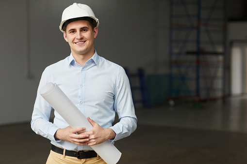 Portrait of young architect in work helmet holding blueprint and smiling at camera while standing in empty building