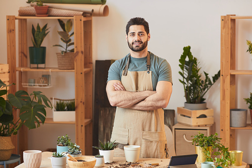 Waist up portrait of handsome bearded man wearing apron and looking at camera while standing by table with houseplants and gardening tools, copy space