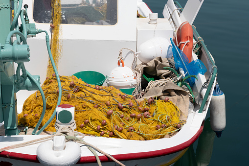 Detail of a fishing boat tied up in a harbor of the Greek Island of Chios in the Aegean Sea. Fishing nets of yellow nylon and floating lines (with small red plastic floats), as well as other fishing gear are lying on the deck of its bow on a sunny day in summer. The fishermen places their nets in the sea at the evening as it gets dark and return to collect the nets with their catch of fish the next morning at sunrise.