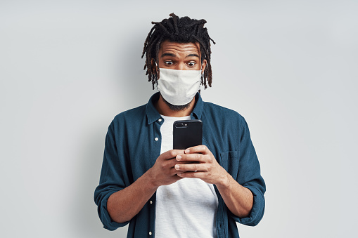 Surprised young African man wearing medical face mask and using smart phone while standing against grey background