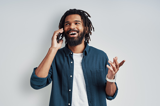 Smiling young African man in casual wear talking on the phone while standing against grey background