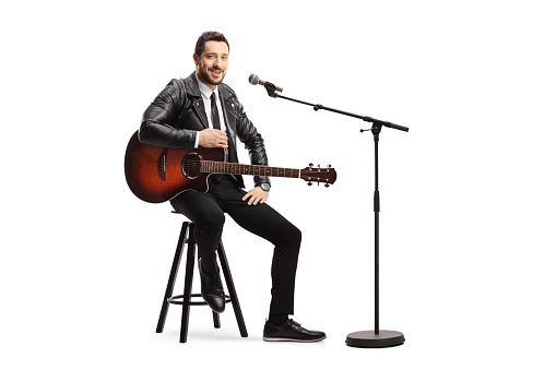 Male singer with an acoustic guitar sitting on a chair with a microphone isolated on white background