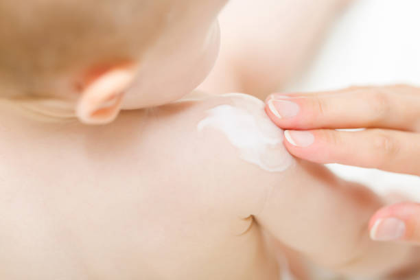 어린 어머니 손가락은 아기 어깨에 흰색 보습 크림을 적용합니다. 깨끗하고 부드러운 몸 피부를 케어하십시오. 근접 촬영. - 연고 뉴스 사진 이미지