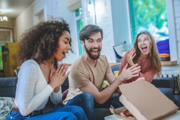 istock Cheerful guy opening box with pizza and two enthusiastic girls 1253484408