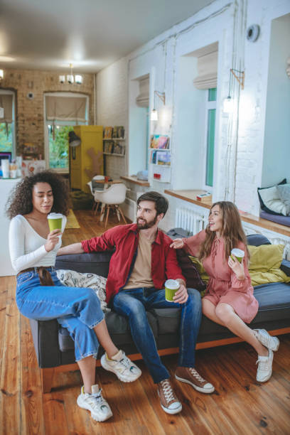 istock Guy and two girls with coffee sitting on sofa 1253484667