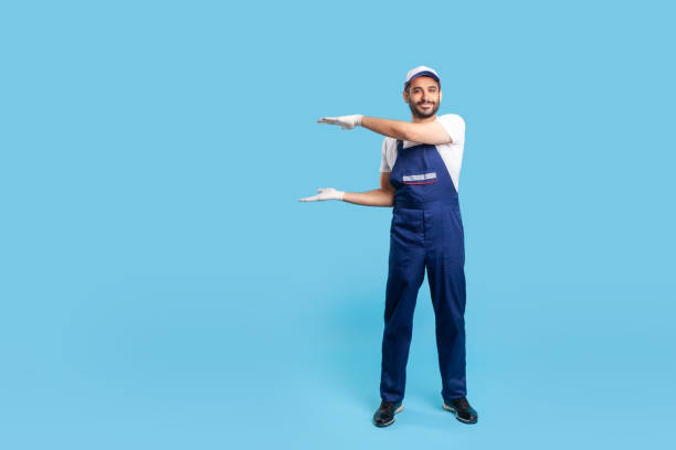 istock Full length happy workman in blue overalls, cap and protective gloves standing, holding empty space 1253484682