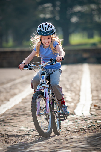 A very happy young blonde girl rides a bike along a cobbled path in golden sunlight
