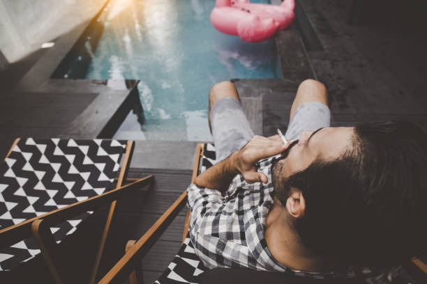 young man smoking cigarette or tobacco near swimming pool. guy addicted cigarette and always smoke a lot of tobacco. he smoke cigarette at hotel. handsome male blowing smoke of cigarette and get relax - nicotine healthcare and medicine smoking issues lifestyles imagens e fotografias de stock