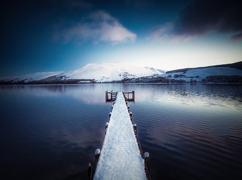 Snow covering a wooden jetty and the hills beyond the shore of Loch Earn in the Trossachs, Scotland. Photographed at dusk in January.