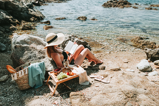 Woman in bikini and hat alone at beach reading electronic book, picnic by the sea outdoors. Young woman enjoying time at mediterranean beach, sunbathing and reading with tray of fruits and wine