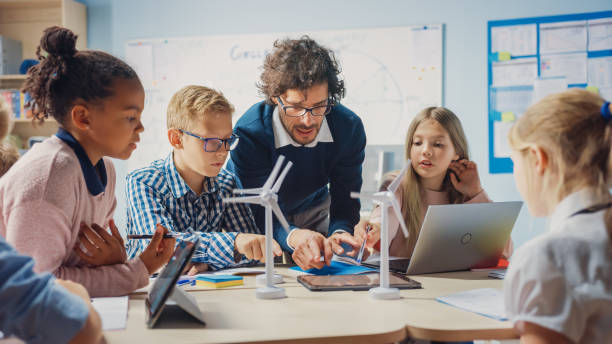 classe d’école primaire : l’enseignant enthousiaste tenant l’ordinateur de tablette explique à un jeune enfant brillant comment les éoliennes fonctionnent. enfants apprenant sur les formes écologiques d’énergie renouvelable - teacher child student education photos et images de collection