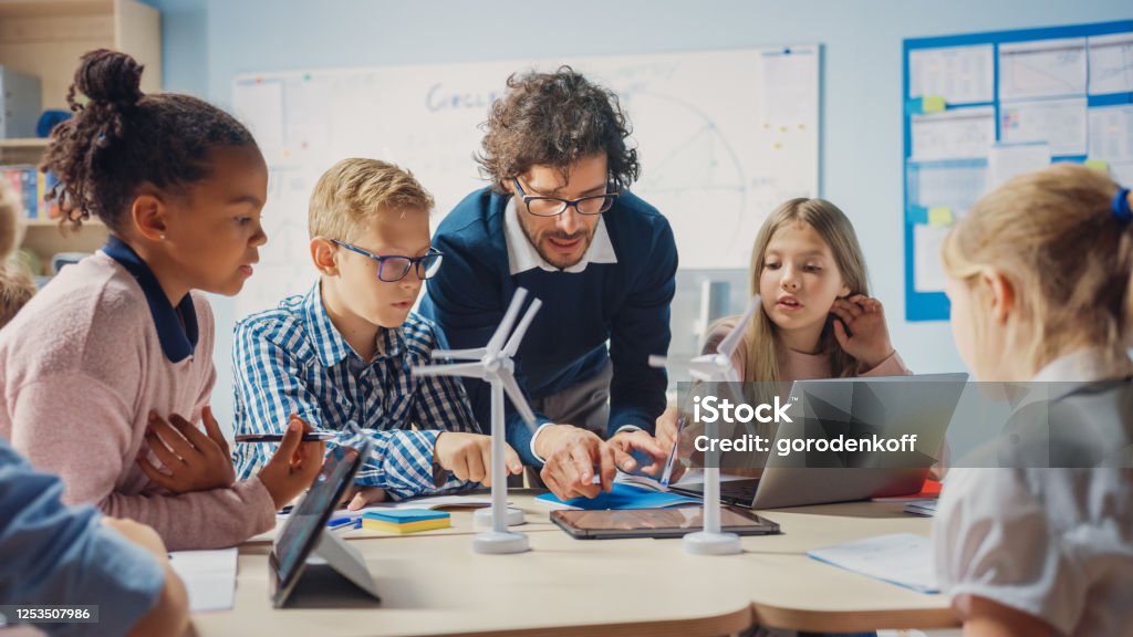 Elementary School Classroom: Enthusiastic Teacher Holding Tablet Computer Explains to a Brilliant Young Children How Wind Turbines Work. Kids Learning about Eco-Friendly Forms of Renewable Energy Teacher Stock Photo