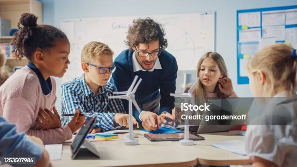 Elementary School Classroom Enthusiastic Teacher Holding Tablet Computer Explains To A Brilliant Young Children How Wind Turbines Work Kids Learning About Ecofriendly Forms Of Renewable Energy Stock Photo - Download Image Now
