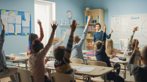 istock Caring Teacher Explains Lesson to a Classroom Full of Bright Diverse Children. In Elementary School with Group of Smart Multiethnic Kids Learning Science, Whole Classroom Raising Hands Knowing Answer 1253508059