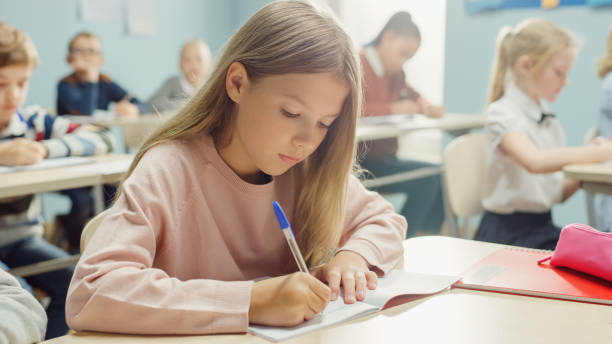 istock In Elementary School Classroom Caucasian Girl Writes in Exercise Notebook, Taking Test and Writing Exam. Junior Classroom with Diverse Group of Children Working Diligently and Learning New Stuff 1253508074