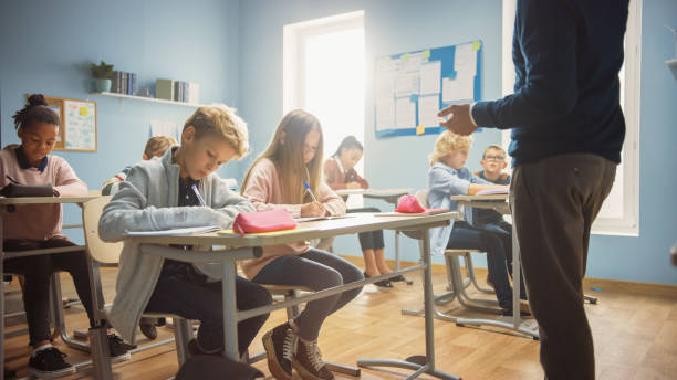 istock Enthusiastic Teacher Explains Lesson to a Classroom Full of Diverse Children. In Elementary School Group of Smart Multiethnic Kids Writing in Exercise Notebooks and Learning. 1253508094