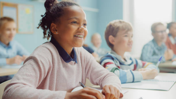 in der grundschulklasse: porträt eines brillanten schwarzen mädchens mit zahnspangen schreibt in übung notebook, lächelt. junior classroom mit diversifizierter gruppe von kindern, die neue sachen lernen - pre teen boy stock-fotos und bilder