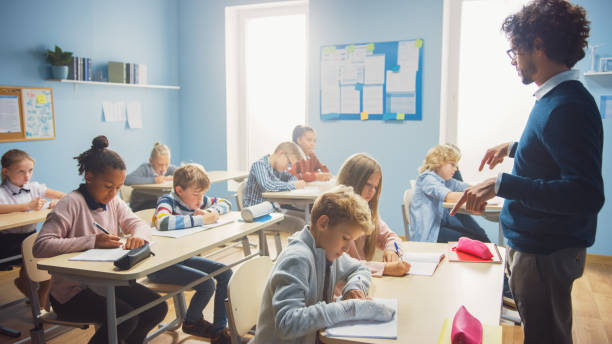 istock In Elementary School Class: Enthusiastic Teacher walks between Rows of Bright Diverse Children, Dictates Lesson. Group of Smart Multiethnic Kids Writing in Exercise Notebooks. 1253508143