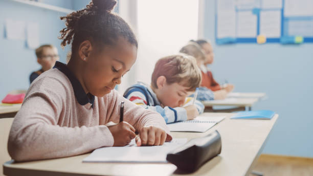 in der grundschule klassenzimmer brillante schwarze mädchen schreibt in übung notebook, nehmen test. junior classroom mit verschiedenen gruppe von hellen kindern arbeiten fleißig und lernen. seitenansicht portrait - junior school stock-fotos und bilder