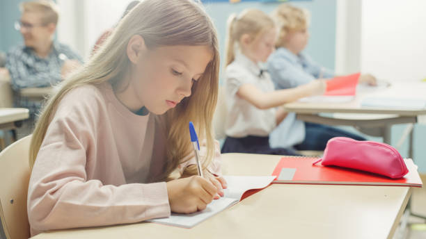 istock In Elementary School Classroom Caucasian Girl Writes in Exercise Notebook, Taking Test and Writing Exam. Junior Classroom with Diverse Group of Children Working Diligently and Learning New Stuff 1253508159
