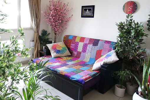 Stock photo showing a lounge sofa covered with a brightly, multicoloured patchwork quilt and patterned scatter cushions. The lounge has a white tiled floor and contains plant pots full of a variety of plants.