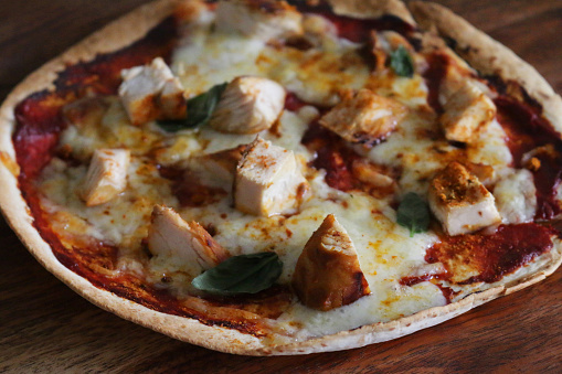 Stock photo showing close-up view of homemade pizza recipe just baked in hot oven made on flour tortilla base, with diced chicken, bubbling stringy mozzarella cheese and Italian family tomato sauce recipe for topping and fresh basil leaves herbs.