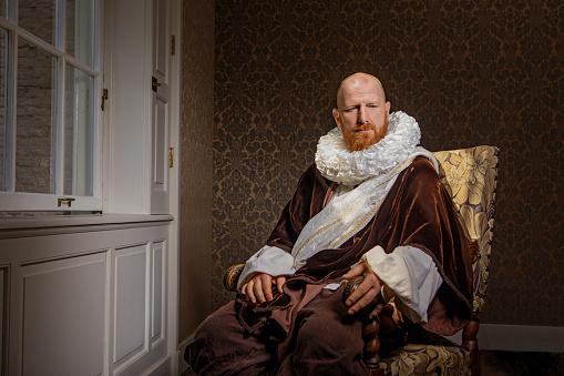 Handsome redhead traditional dutch man wearing historically correct outfit reading a book by candlelight in a typical townhouse drawing room