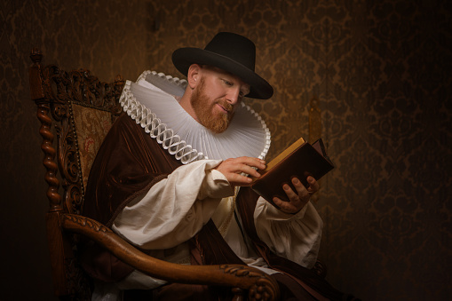 Handsome redhead traditional dutch man wearing historically correct outfit reading a book in a typical townhouse drawing room