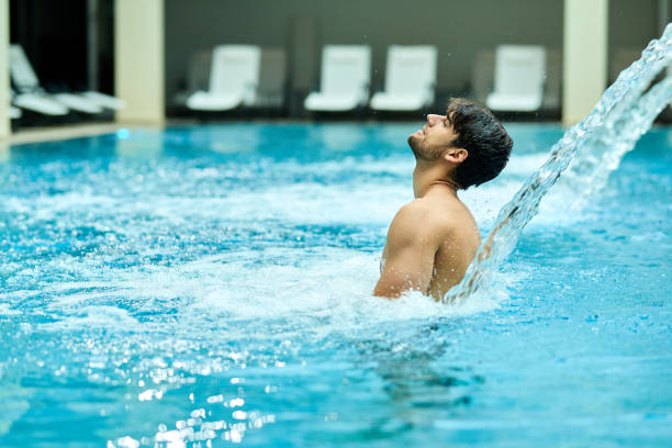 Young man enjoying in hydrotherapy at health spa. Young man spending a day at the spa and relaxing under a stream of water in the swimming pool. thermal pool stock pictures, royalty-free photos & images