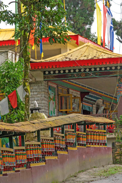 Colorful prayer wheels lining a street in Sikkim India Row of Buddhist prayer wheels leading to a monastery with colorful prayer flags and saffron colored roof on a quiet street buddhist prayer wheel stock pictures, royalty-free photos & images