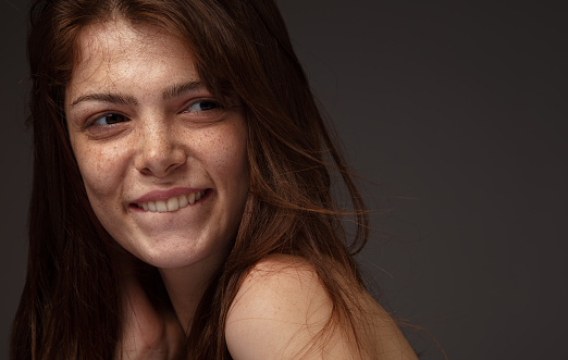 Side view of freckled beautiful young woman smiling and biting her lip front of grey background.