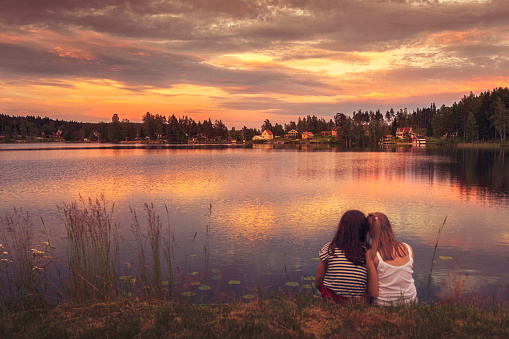 Two girls sitting by the edge of a lake and looking towards the evening sunset in Dalarna, Sweden.