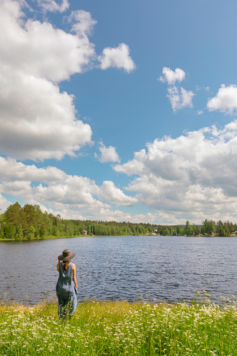 A woman standing at a meadow next to a lake and looks out over the water. Dalarna region, Sweden.