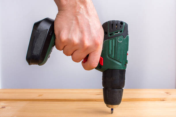 istock Men's hand holding battery drill and drilling wooden board. 1253554785