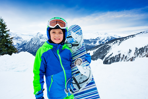 Front portrait of a boy hold snowboard in hands over high mountain peaks panorama